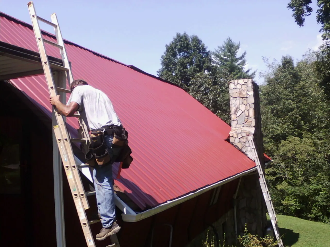 Roofer on a ladder beside a red metal roof and stone chimney — Soffit & Fascia Repair by Roofing Brothers in Allport